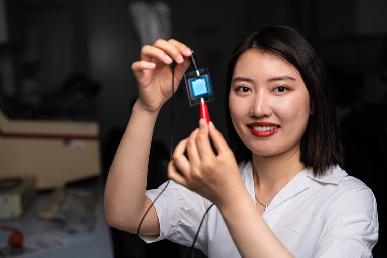 Junge Frau im Labor hält ein kleines technisches Bauteil in der Hand und lächelt.